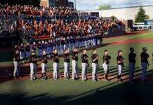 College Pitcher Suffers Serious Brain Injury In Car Crash Baseball players in purple jerseys and white pants stand in a line on the field as a large crowd watches from orange-seated stands behind them.