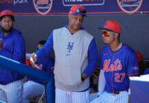 Mets Legend Sends Clear Message to Struggling Superstars Baseball players in Mets uniforms stand in the dugout, one gesturing while another wears orange cap and reflective sunglasses, behind a blue padded railing.