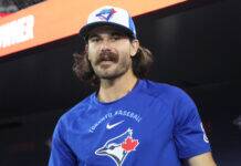 MLB Star Goes Viral For Embarrassing Blunder With Assistant Man with long hair and a mustache wearing a blue Toronto baseball shirt and a Toronto Blue Jays cap, standing in a dark setting with a red banner behind him.