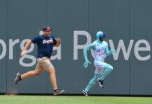 Atlanta Braves Fan Taunts Popular Mascot Two men sprinting side by side along a ballpark outfield wall; one wears a navy Braves shirt, beige shorts, and a cap, the other in a turquoise bodysuit with goggles.
