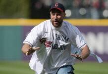 Travis Kelce Buys Massive Stake In His Beloved MLB Team Man in a white baseball jersey and jeans running toward the camera on a sunny field, wearing a dark cap with a red brim.