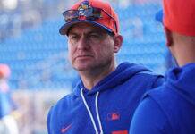 Mets Manager Calls Out Team With Season Slipping Away Portrait of a man in a blue hoodie at a baseball stadium, orange cap with sunglasses perched on the brim, blue seats in the background kept out of focus.