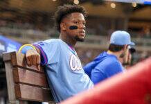 The Braves Pressure is Mounting as Injured List Grows Baseball player in a light-blue jersey sits on a wooden dugout bench with eye-black under his eyes, stadium lights blurred in the background.